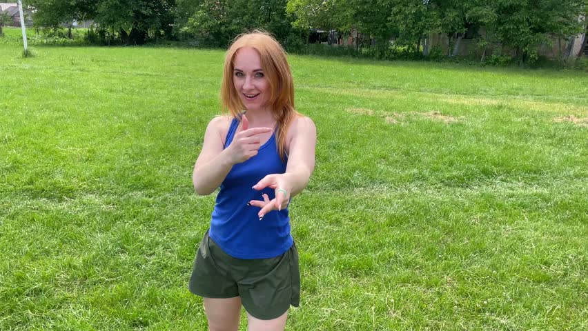 Young woman enjoys dancing alone in a sunny park, surrounded by grass and trees, embracing a healthy lifestyle