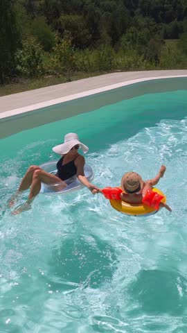 A funny little boy, wearing sunglasses, a hat, and arm floaties, is swimming in the pool with inflatable rings along with his mom on a sunny day, enjoying summer together. Vertical video