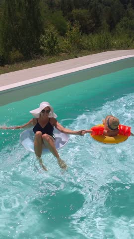 A funny little boy, wearing sunglasses, a hat, and arm floaties, is swimming in the pool with inflatable rings along with his mom on a sunny day, enjoying summer together. Vertical video