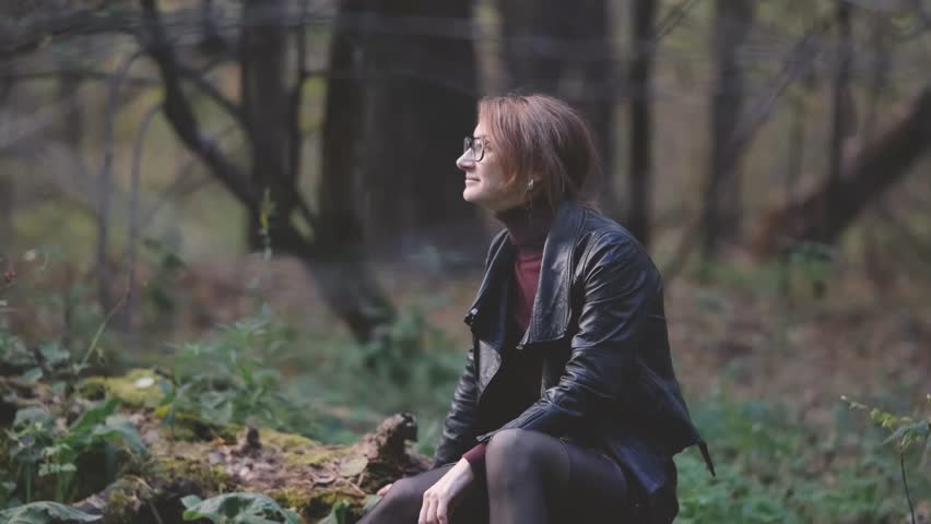 Smiling city woman relaxing in the forest. Thoughtful woman in glasses and office clothes sitting casually on an old fallen tree covered with moss outdoors, breathing fresh air, enjoying nature.