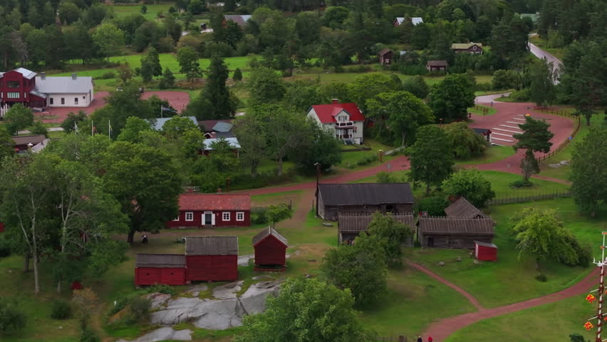 Lush green trees surround a quaint village with traditional red and white Finnish buildings and a gravel road winding through