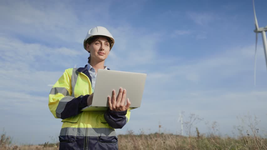 View from below of charming Caucasian woman standing still and looking forward. Holding laptop with one hand. Inspecting wind turbines in middle of power plant. Recording observations. Green energy.