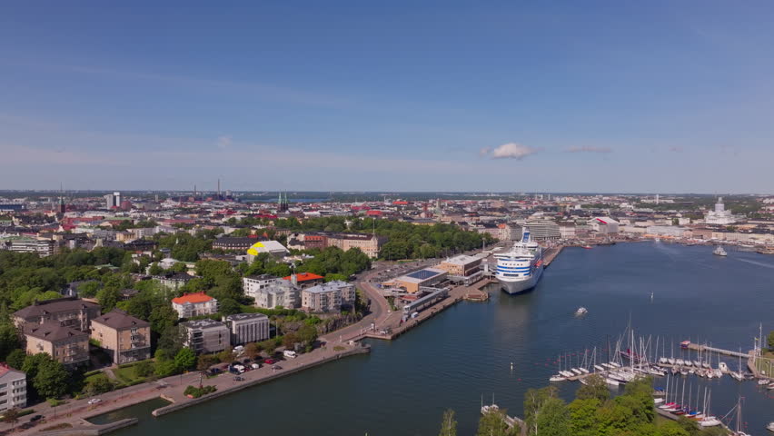 Aerial views of Helsinki, Finland capture a large cruise ship docking on the harbor on a sunny summer day, showcasing the lively cityscape and bustling waterfront activities