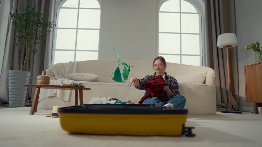Girl throwing bikini and clothing into big suitcase, preparing to vacation trip. Happiness and joy of future travel in summer holidays, young woman packing suitcase at home, carefree female tourist