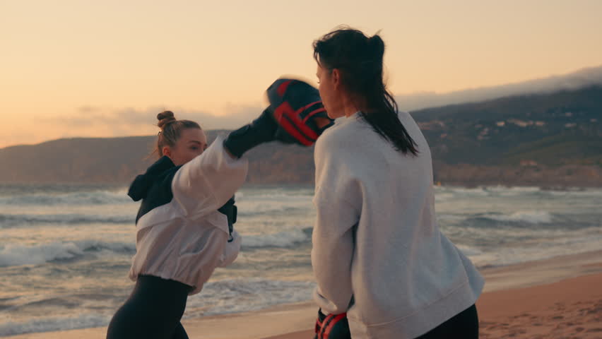 Determined female fighters practicing defence boxing at beach during sunset. Strong young women boxers are exercising in nature. 