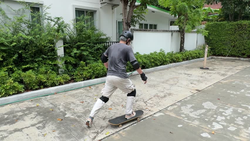 The senior Asian man is trying to ride a skateboard in the village during the daytime