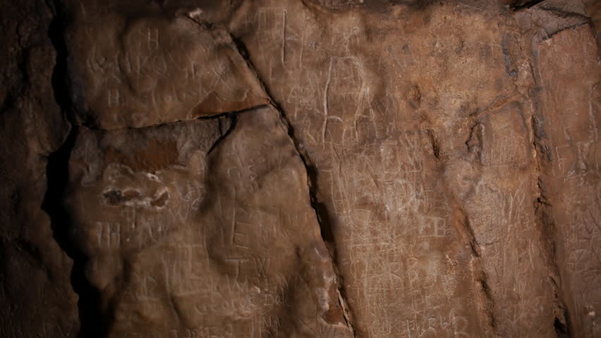 A cave wall with carvings, runes and inscriptions etched into the stone. The uneven surface and dim lighting add to the ancient and mysterious atmosphere. Marks left by past visitors and explorers