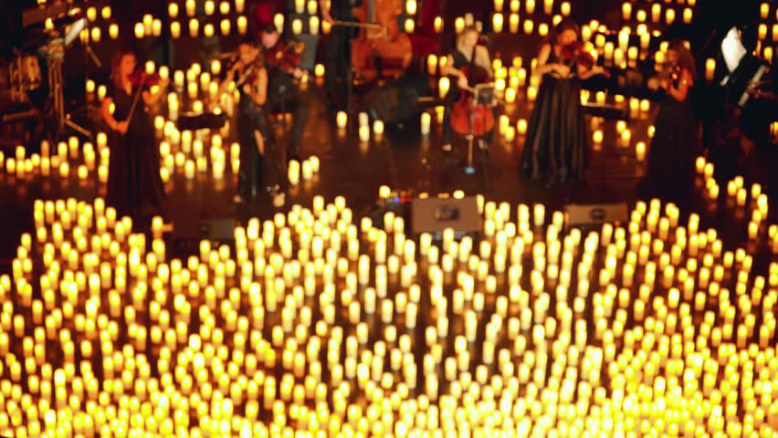 An orchestra with various instruments performs on a dimly lit concert stage, illuminated by many candles. Blurred video