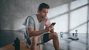 Sweaty man swiping smartphone relaxing at loft location zoom on. Professional tired powerlifter resting at bench after cardio training workout. Calm athlete sitting at empty gym looking mobile phone - Powered by Shutterstock - Get 15% off with code: PIKWIZARD15