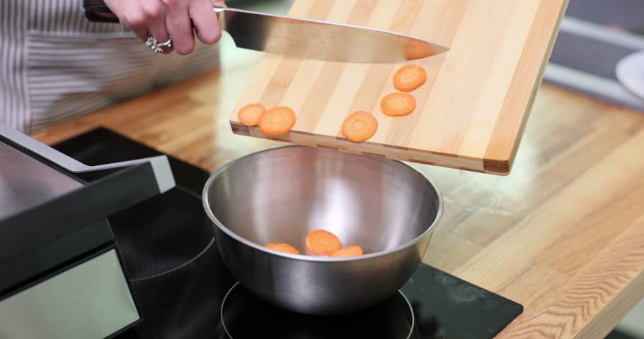 Housewife pours chopped carrot into metal bowl from cutting board using knife in kitchen. Woman prepares healthy ingredient for cooking meal slow motion