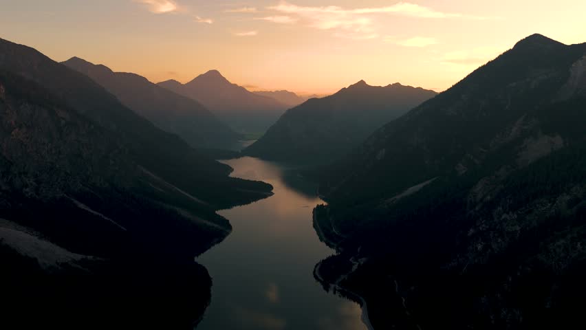 Witness the breathtaking transition as the sun sets behind majestic mountains, casting warm golden tones on the tranquil lake below. A peaceful moment that captures nature