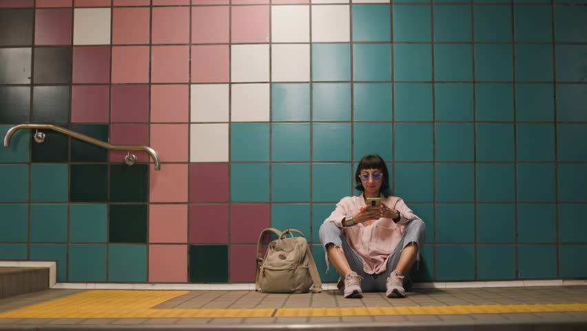 Young woman waiting for train at station and using her phone. watch schedule. Traveling by public transport.  Young girl in stylish clothes with backpack in subway.  - Powered by Shutterstock - Get 15% off with code: PIKWIZARD15