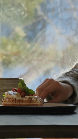 Vertical video. A young woman enjoys a sweet breakfast waffle dessert and tea while seated in a cozy cafe surrounded by a serene winter forest landscape