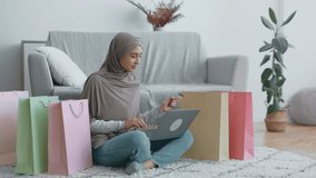 A woman in a gray hijab browses online for products using her laptop, surrounded by vibrant shopping bags in a modern living space. - Powered by Shutterstock - Get 15% off with code: PIKWIZARD15