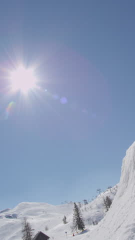 SLOW MOTION VERTICAL: Pro snowboarder jumps upside down and does a handplant trick over sun in snow park. Guy snowboarding and jumping in terrain park in sunny ski resort. Snowboarder shredding park