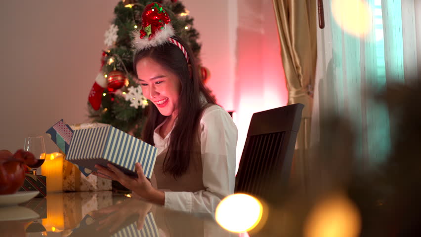 Happy beautiful Asian woman is excited to open or unbox the Christmas presents in Christmas Eve night party. Excited Asian woman is opening a Christmas's gift box with emotional expression.