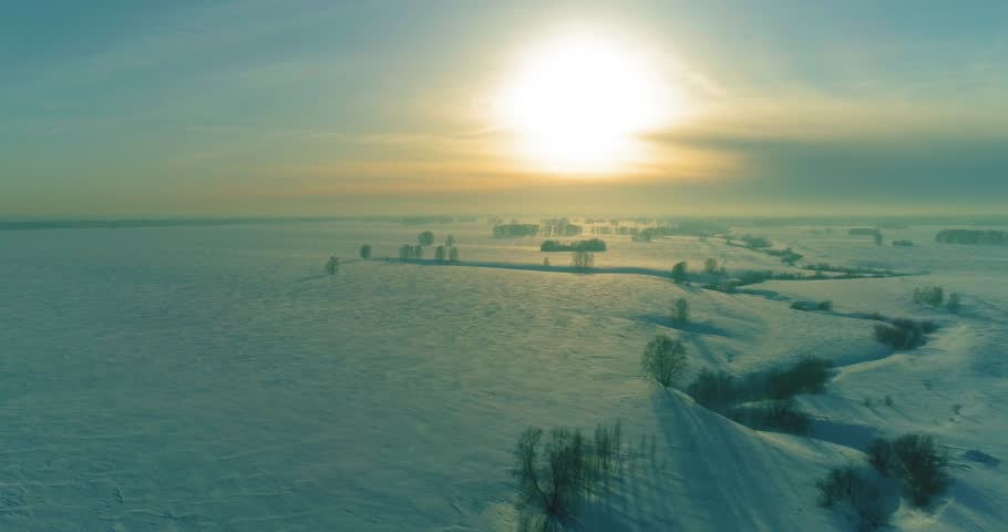 Drone aerial view of cold winter landscape arctic field, trees covered with frost snow, ice river and sun rays over horizon. Extreme low temperature weather.