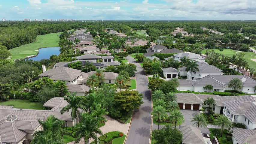 Residential neighborhood with luxury homes and lush greenery. Palm trees line the streets as the camera flies above modern properties. Golf greens and water features stretch in the distance. Aerial.