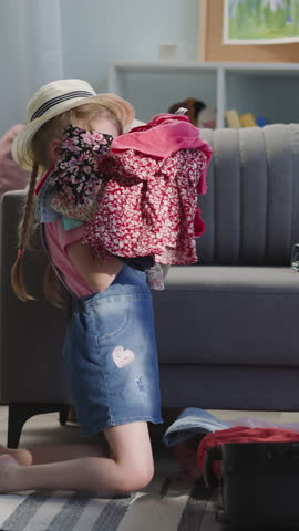 Joyful preschooler girl with panama puts clothes into bag and imitates flying airliner at home. Playful child dreams about travel to foreign country