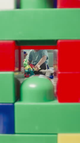Children play with baby doll and airplane sitting near wooden house. Girl and boy spend time in nursery view through clearance in construction of cubes