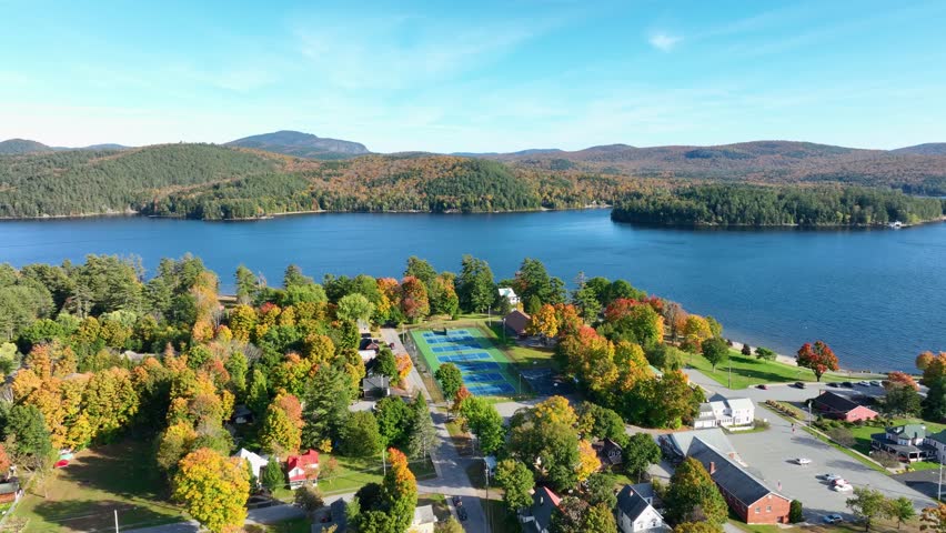 Aerial view of Schroon Lake, New York, showcasing the calm lake, fall foliage, and small town structures nestled along the shoreline under a clear sky.