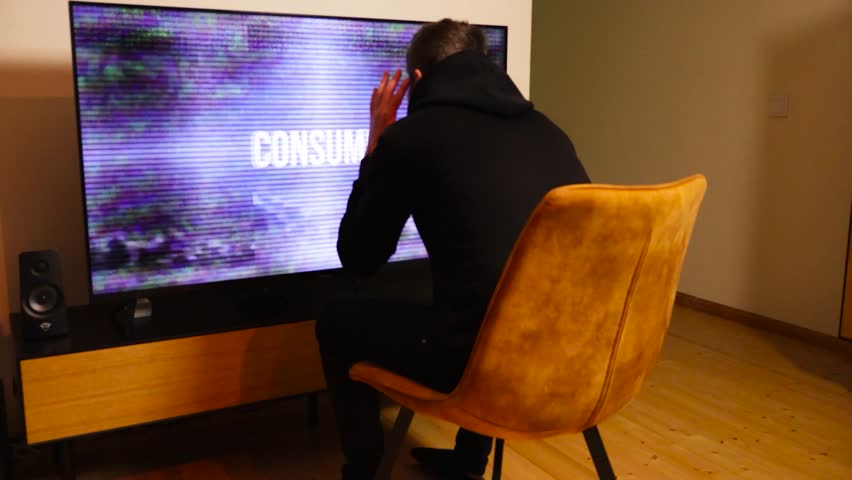 Footage of a man in black clothing sitting in front of a big screen television that shows propaganda messages and is brainwashing him. The man is moving around showing that he is in stress, dark room.