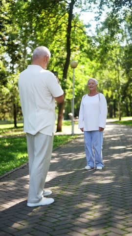 Vertical shot of elderly gray-haired man taking picture of senior woman posing in sunny park on paved path. Happy mature aged wife and husband enjoying free time on summer sunny day, slow motion.