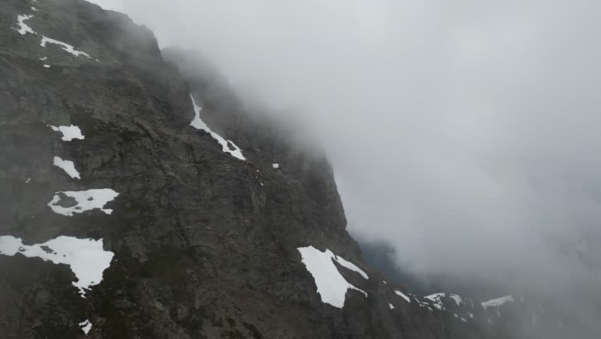 Aerial view of a steep, rocky mountain peak shrouded in thick fog, with patches of snow clinging to its slopes, evoking a sense of mystery and awe. British Columbia, Canada.