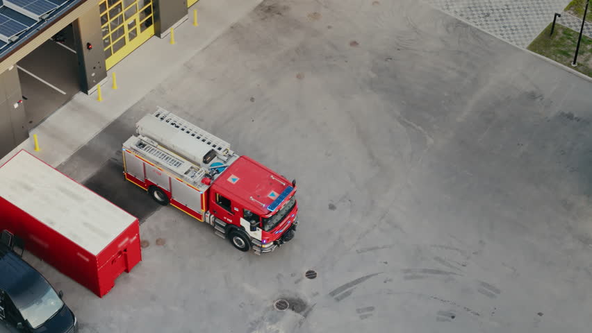 Aerial Drone Footage of a Fire Truck with a Team of First Responders Drives Out with Lights Flashing. It Races Onto the Street, Responding to an Emergency Situation, Ready to Save Lives