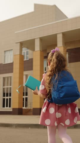 Happy cute little girl with backpack and book running to elementary school class outdoor back view. Adorable female kid child pupil enjoy primary education hurry to classroom lesson learning studying