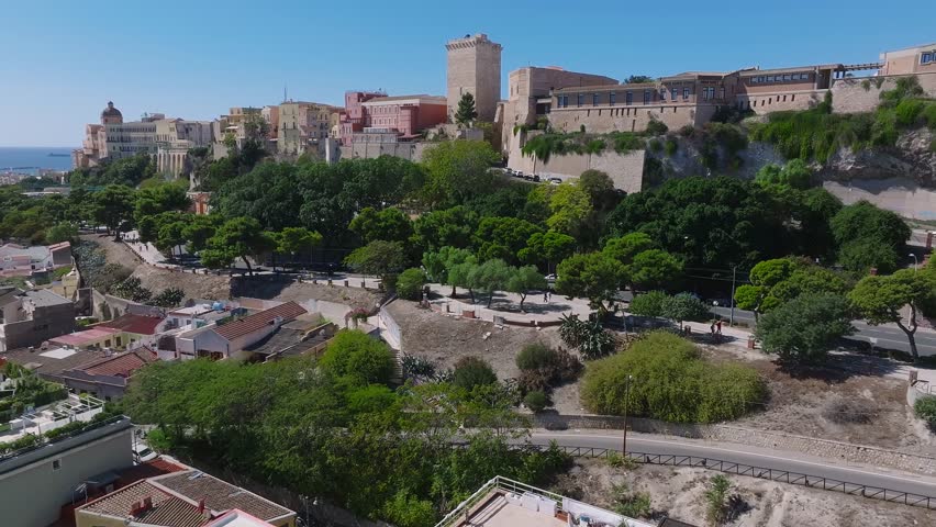 Aerial view of a historic fortress on a rocky hill in Cagliari, surrounded by greenery and urban buildings, with a bustling roundabout and clear blue sky.