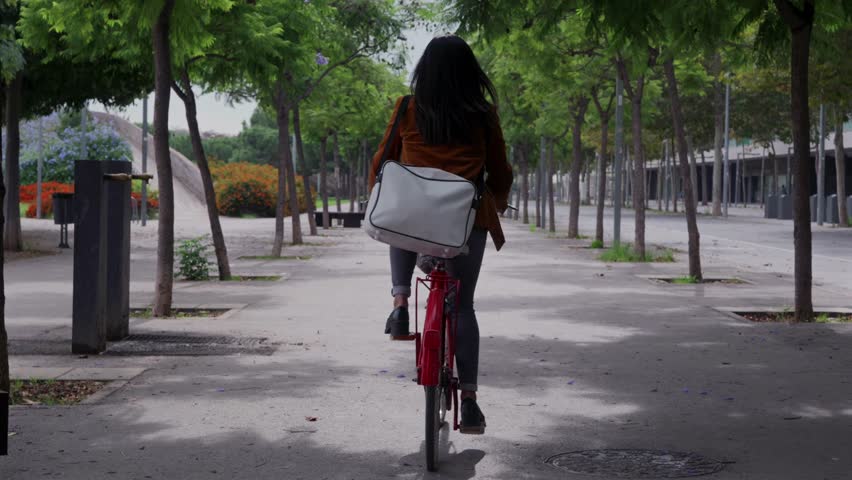 A woman dressed in formal clothing rides to work on a red bicycle passing through a tree-lined street.