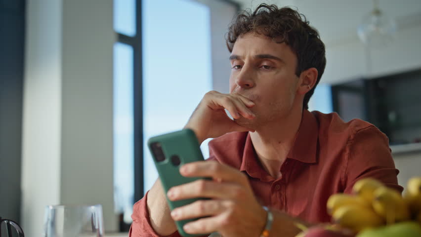 Frustrated man looking cellphone sitting apartment kitchen alone closeup. Tired guy reading bad news at smartphone feeling exhausted at home. Stressed male disappointed mobile phone message at morning