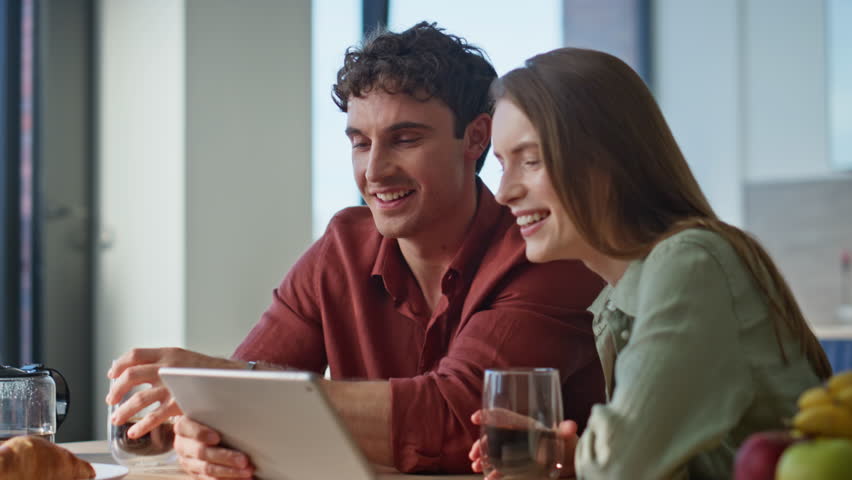 Pair watching tablet video holding coffee cups closeup. Smiling family couple enjoy home breakfast laughing on online content sitting kitchen. Happy soulmates looking pad computer in modern apartment.