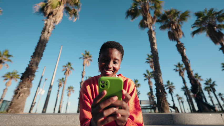 Smiling Black woman in bright sweater looking at smartphone while sitting outdoors with palm trees in background. Engaged and relaxed, she scrolls through social media on cellphone device application