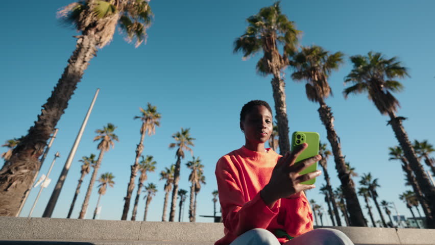Young African American woman in a bright sweater gesturing during video call on smartphone. Female technology user engages in conversation, surrounded by palm trees. communication and 5G internet