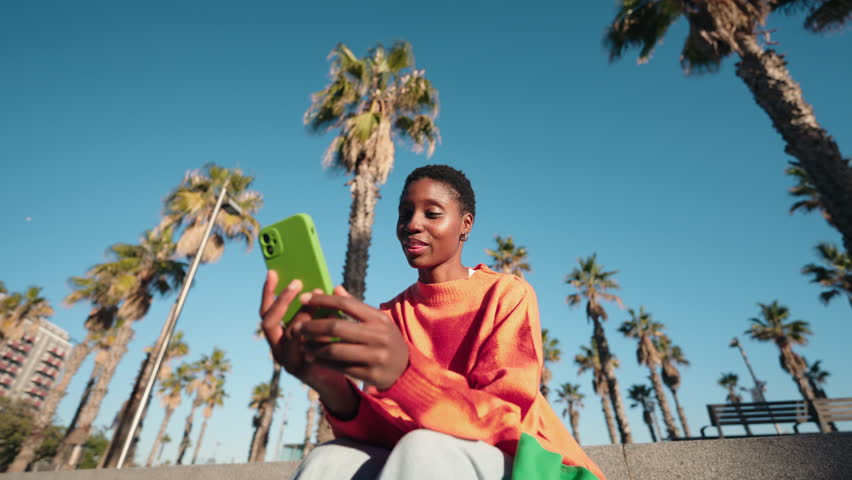 Young African American woman in a bright sweater gesturing during video call on smartphone. Female technology user engages in conversation, surrounded by palm trees. communication and 5G internet