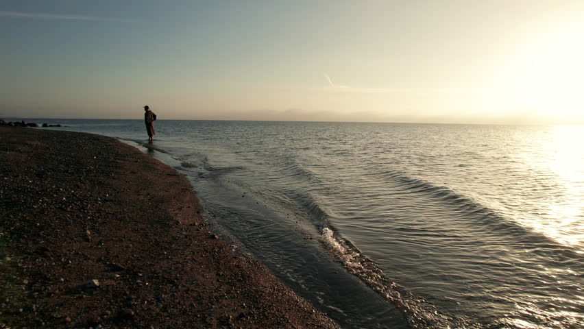Man strolling along a serene seashore, with gentle waves lapping at the shore during sunrise