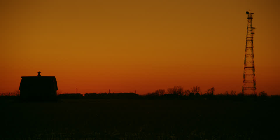 Lone country barn and a communications tower share a field and a blazing orange sky during the golden hour after sunset. 