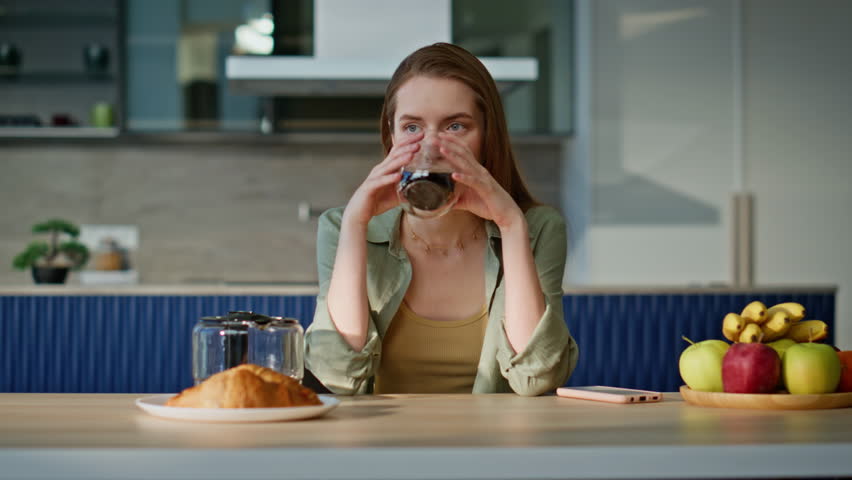 Peaceful woman enjoying coffee sitting apartment kitchen alone closeup. Relaxed beautiful lady drinking aromatic beverage from glass cup having home breakfast. Calm young girl start morning with smile