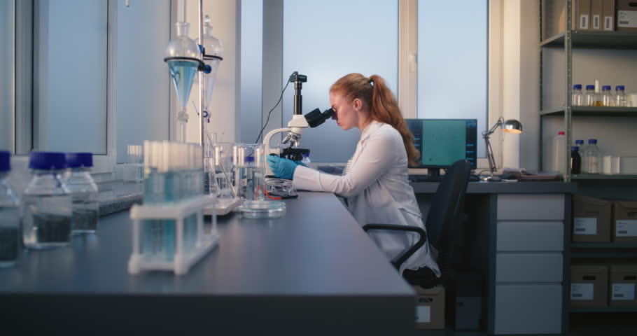 Young female scientist sits in chair, looks under microscope and examines samples of experimental drugs in medical science laboratory. Innovations in pharmaceutics and pharmacology. Medicine concept.