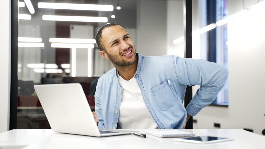 Sick tired African American businessman suffering from back pain while working on laptop sitting at desk at workplace in business office. Upset young adult worker massages painful lower back muscles