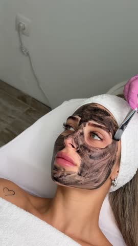 cosmetologist applying black mask on pretty woman face wearing black gloves, gorgeous woman in spa having facial procedures