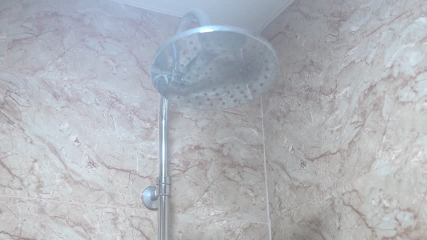 Water is pouring from round shower head in bathroom. Steam is coming up from hot water. Wall is covered in marble-like tiles