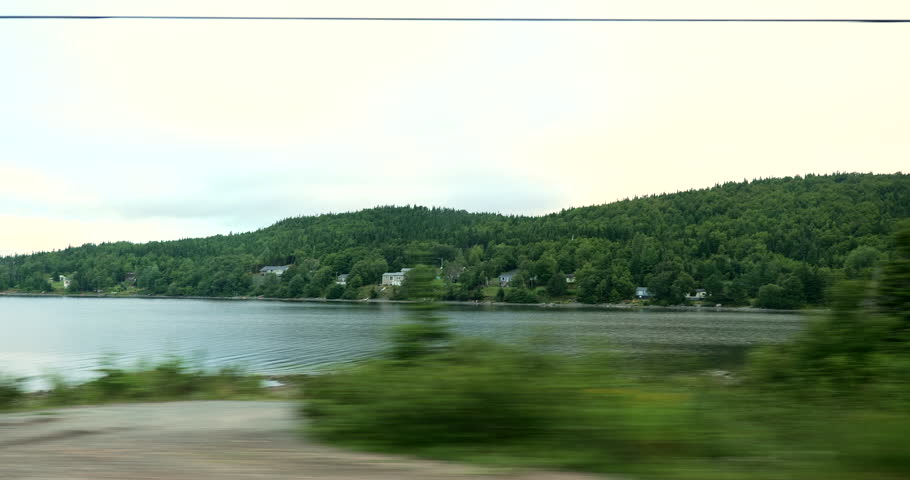 Side driving plate on a highway through a forested area and lake in Ontario, Canada. Road surrounded by dense trees under partly cloudy sky. Forest travel and nature exploration concept