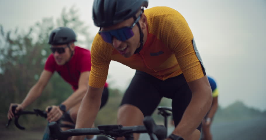 Close Up Portrait of a Cyclist in an Orange and Yellow Jersey Racing on a Misty Road Against Competitors. Intense Dynamic Focus and Pedaling Highlight the Thrill of Cycling Outdoors