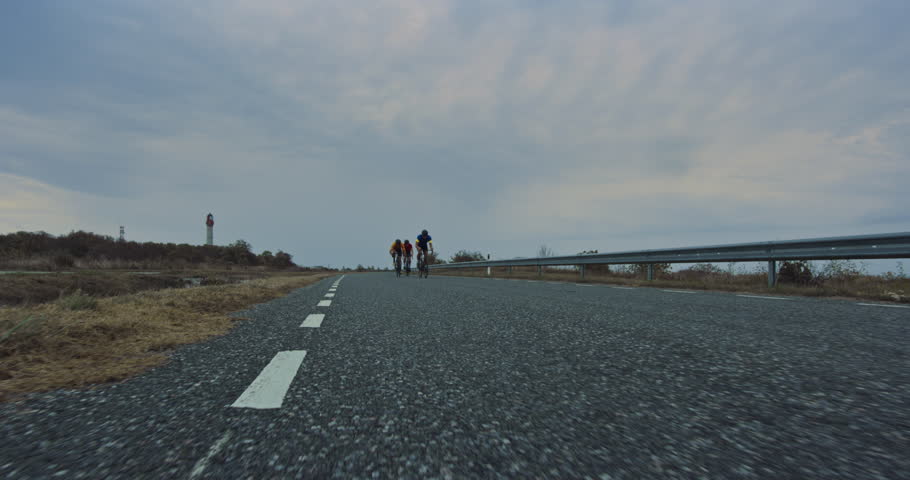 Three Athletes Ride Together Along the Road, Pedaling Bicycles as They Practice on a Country Road. Sportsman in Blue Jersey Leading the Pack and Providing Clean Air. Low Angle Handheld Footage