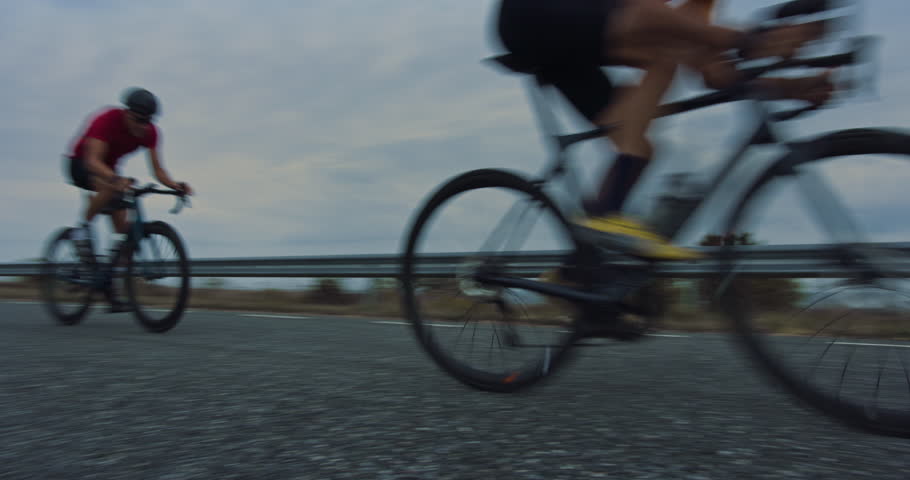 Three Athletes Ride Together Along the Road, Pedaling Bicycles as They Practice on a Country Road. Sportsman in Blue Jersey Leading the Pack and Providing Clean Air. Low Angle Handheld Footage
