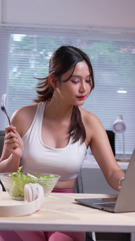 Young woman in sportswear enjoys a salad while working on her laptop at home, balancing nutrition and productivity