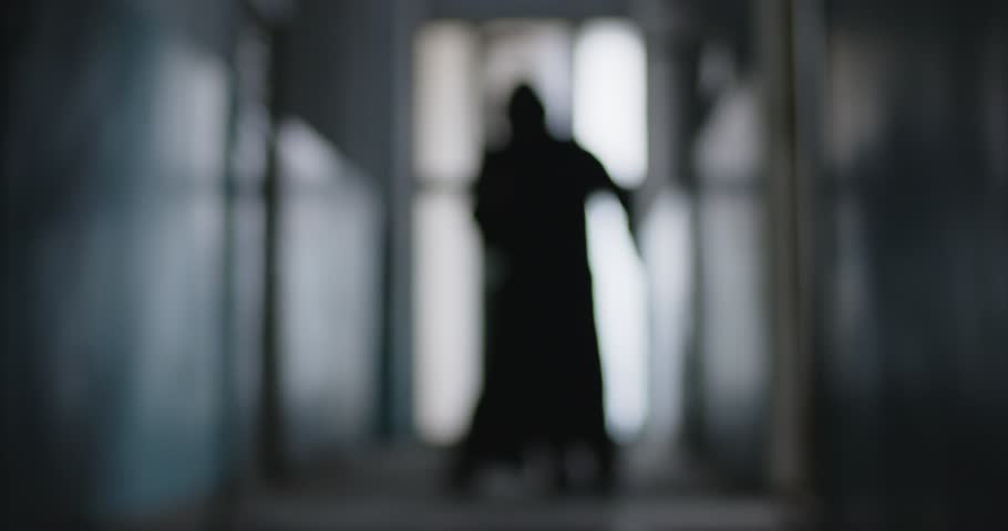 Blurred defocused silhouette of person in grim reaper death costume in black cloak holding scythe walking indoors on dark background. Halloween and horror concept. 4k video. Slow motion video.